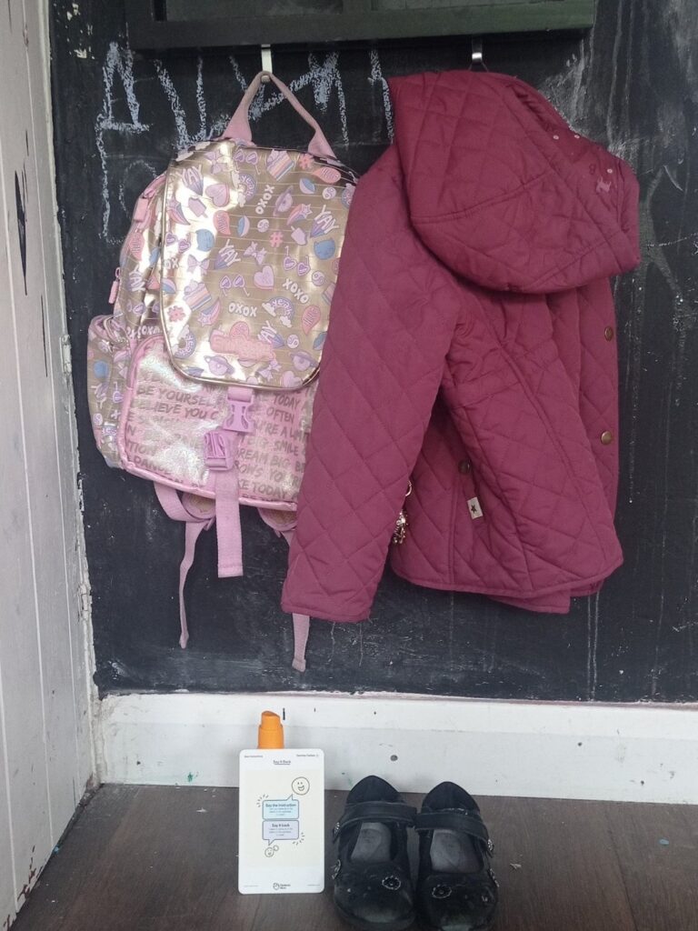 A school bag and coat hung on pegs with shoes and a water bottle lined up underneath, ready for the morning